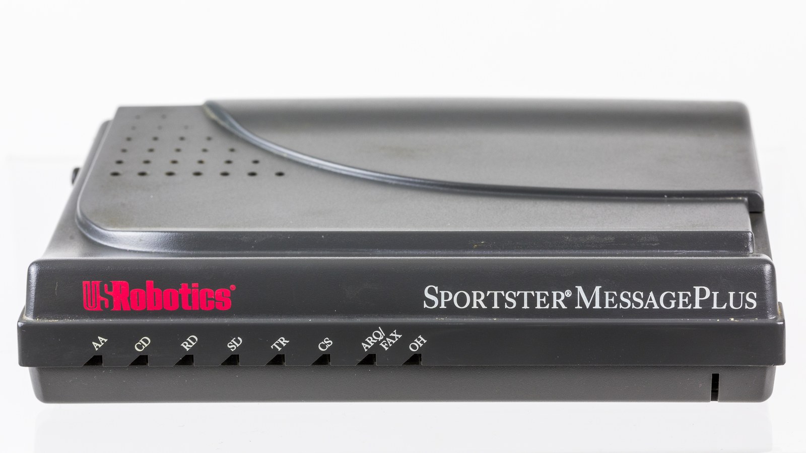 A dark grey external modem photographed against a plain white ground, seen low and slightly from the front. The case is a flat wedge of moulded plastic about the size of a hardback book, with a shallow lip along its front edge carrying a row of eight small indicator windows, each labelled with a two-letter abbreviation. A manufacturer's name is printed at the left of the same edge. No cables are attached.