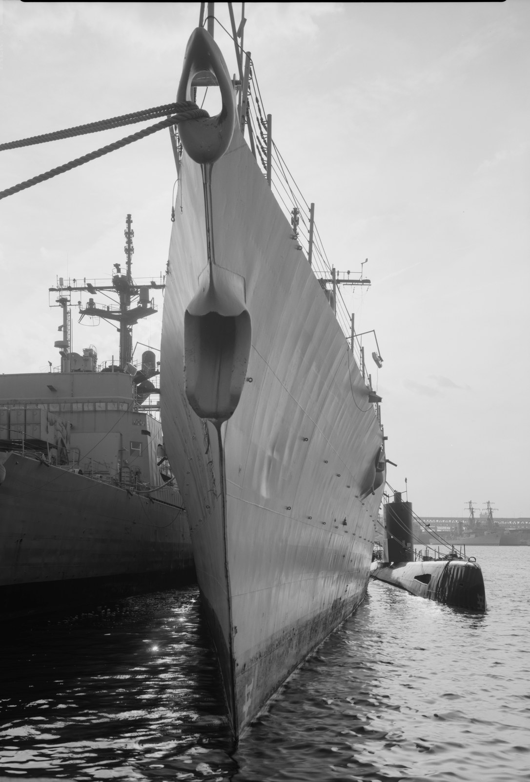 A black-and-white photograph looking along the waterline of a moored warship. The bow of a grey frigate rises steeply and fills the centre of the frame, mooring lines running off to the left, its hull plating rippled with weld seams. A second warship lies alongside to the left. To the right, low in the water, the black hull and sail of a submarine are made fast against a pier. More laid-up ships and cranes are visible in the distance across flat water under a blank sky.