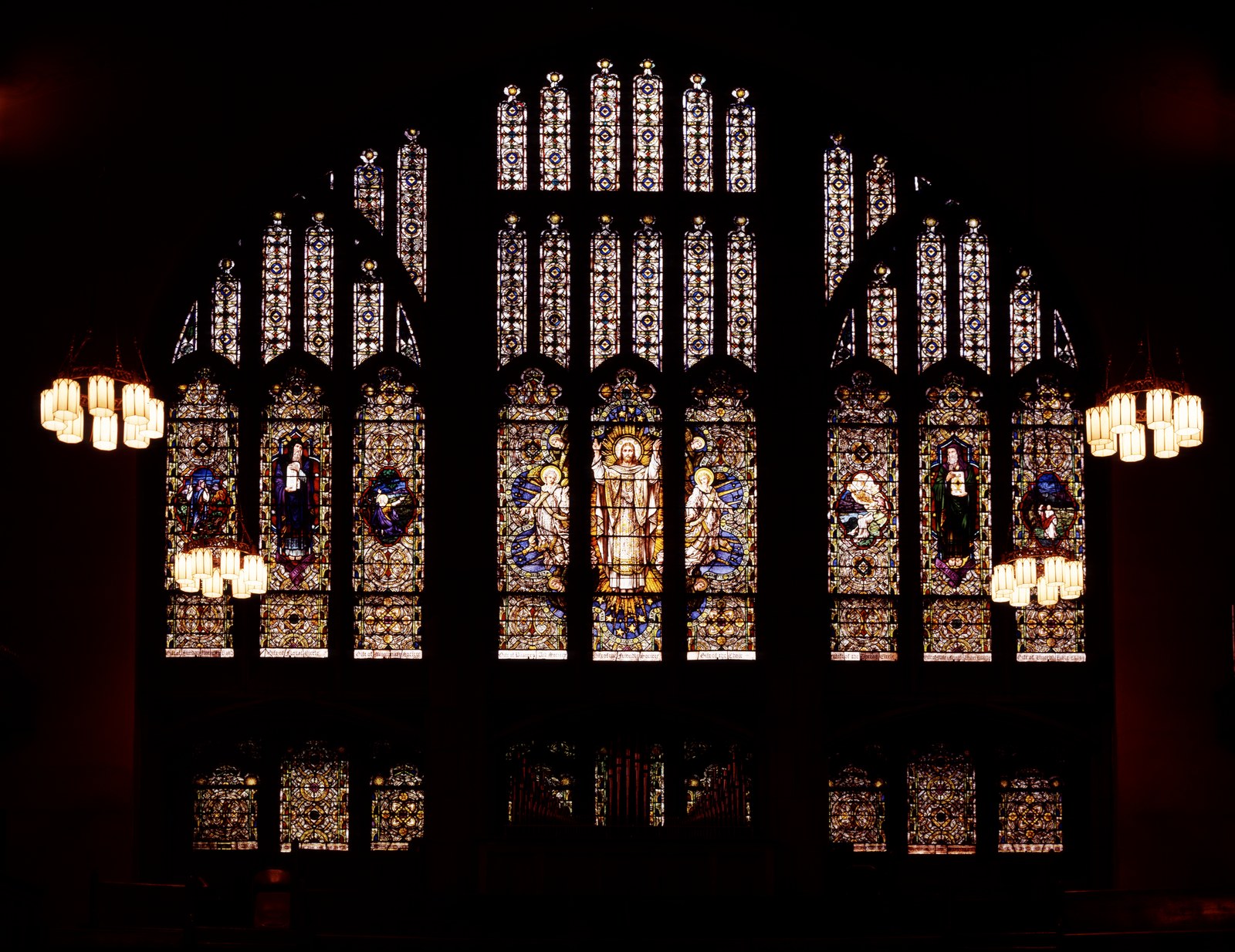 The interior end wall of a church sanctuary photographed in near darkness, so that the lit stained glass carries almost all the light. A wide pointed arch of leaded windows fills the upper wall: three tall central lights showing a robed figure flanked by two attendants, with three narrower figured lights to either side and rows of small patterned lancets above. Two clusters of frosted chandelier globes hang lit at the left and right edges. Below the glass, organ pipes stand in shadow.
