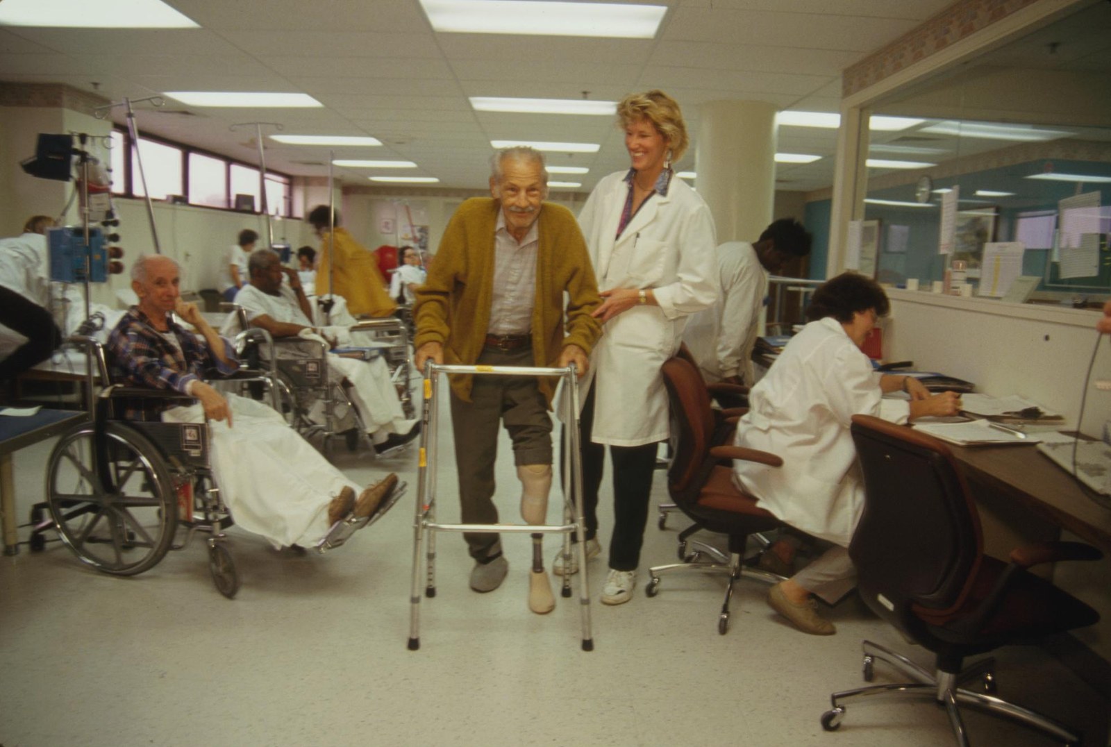Color photograph, fluorescent light, September 1994. An open hospital rehabilitation floor. An elderly man in a mustard cardigan walks toward the camera with an aluminum walker, one leg bandaged below the knee, a prosthetic limb standing on the floor beside him; a physical therapist in a white coat walks at his side with a hand at his back. Behind them two older patients wait in wheelchairs with IV poles and draped sheets, and staff in white coats work at a long counter along the right-hand wall beneath a drop ceiling of fluorescent panels.