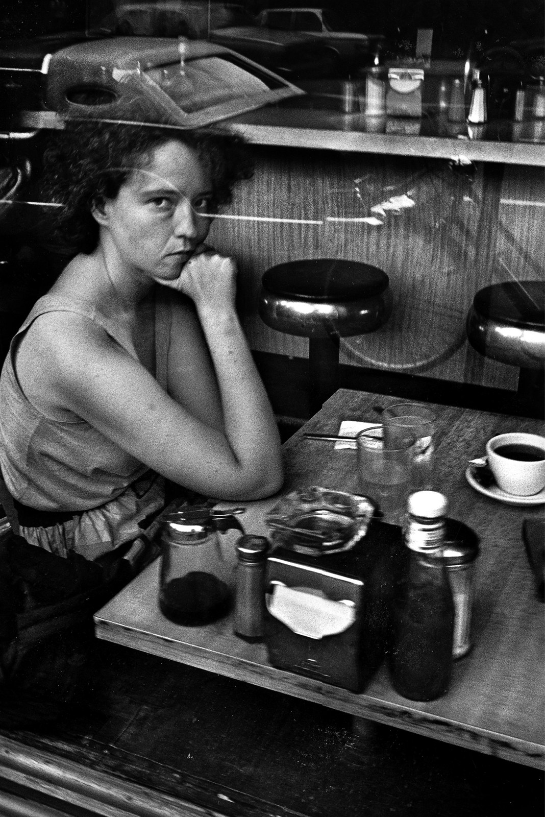 Black-and-white photograph, 1990. Seen through a plate-glass window, a woman sits alone at a wood-topped table in a New York coffee shop, chin on her hand, looking out at the camera. On the table in front of her: a cup of coffee on a saucer, a glass ashtray, a napkin dispenser, salt and pepper shakers, a glass of water. Behind her a row of chrome-stemmed counter stools stands empty along a wood-panelled counter, and parked cars are reflected in the glass above her.