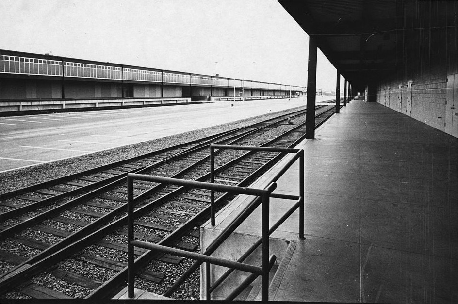 Black-and-white photograph of the Hunts Point produce terminal in the Bronx, empty. A covered concrete unloading platform runs into the distance along a blank block wall on the right; a railroad siding with two sets of track and a low pipe handrail runs beside it; across the tracks, the long low market building with its continuous clerestory window band and a wide empty truck apron marked out in parking bays.