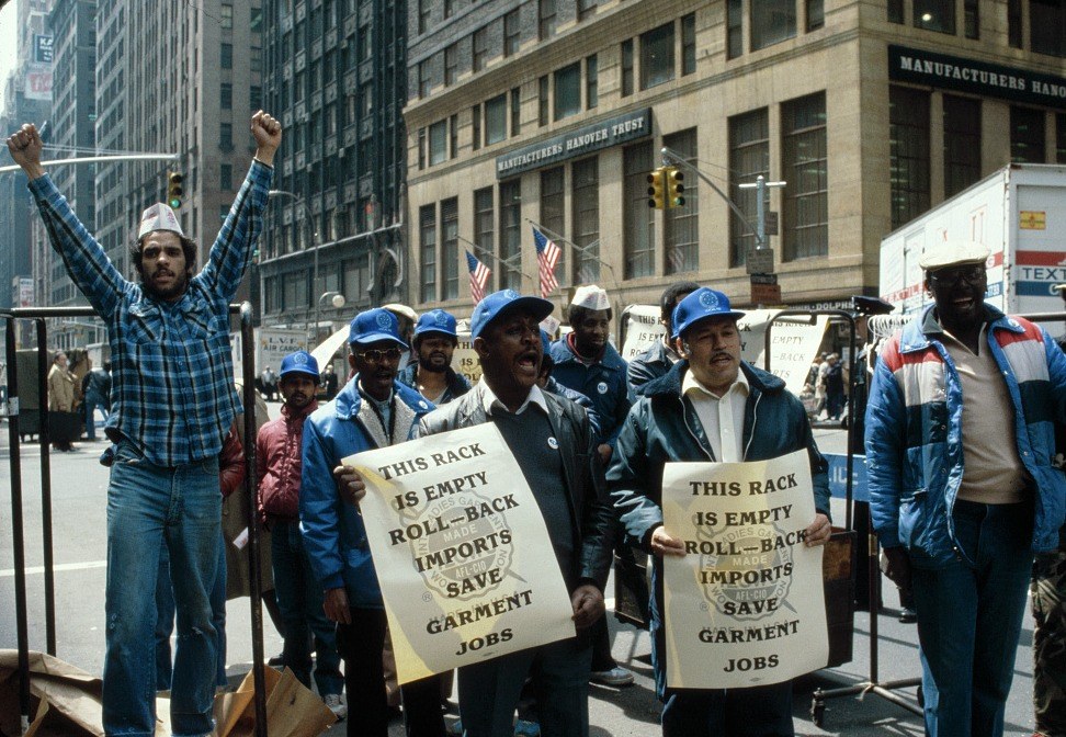 Color photograph of a daytime demonstration on a Midtown Manhattan avenue. A dozen garment workers in union caps and winter jackets stand behind empty wheeled clothing racks, several holding placards reading THIS RACK IS EMPTY — ROLL-BACK IMPORTS — SAVE GARMENT JOBS around the ILGWU union label. One man at the left stands on a barricade with both fists raised, shouting. Behind them, the stone facade of a Manufacturers Hanover Trust branch, American flags on poles, a box truck at the curb.
