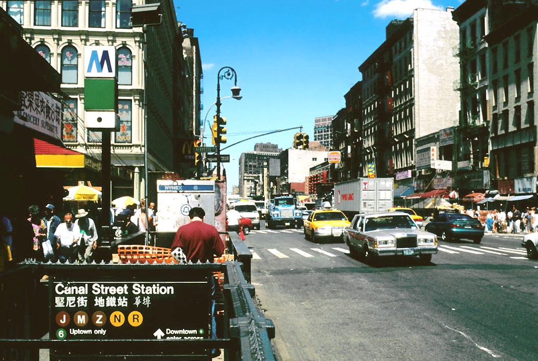 Color photograph looking east along Canal Street in Manhattan on a clear summer day. In the left foreground a subway entrance railing and a vendor bent over a folding table stacked with orange trays; behind him a grocery awning carrying Chinese characters and a green subway lamppost. The roadway carries yellow taxis, a Lincoln Town Car, a box truck and a tractor trailer; tenement blocks with fire escapes and painted signs recede toward the Bowery under a blue sky.