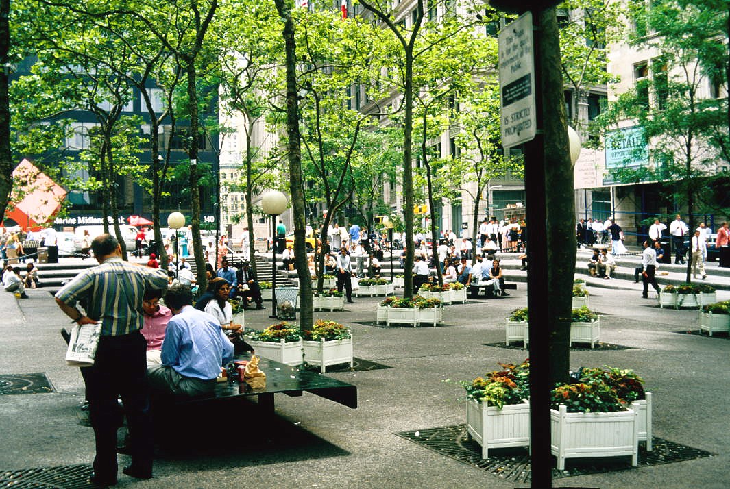 Color photograph of a small downtown Manhattan plaza at midday, seen under plane trees in full leaf. Office workers in shirtsleeves sit on low benches and a steel table eating lunch; one man stands over them holding a folded newspaper and a shopping bag. White wooden planters of flowers are set across the granite paving, and further back dozens more people cross the steps and sidewalk in front of stone office buildings; a bank sign and a retail-leasing banner are visible at the far side.