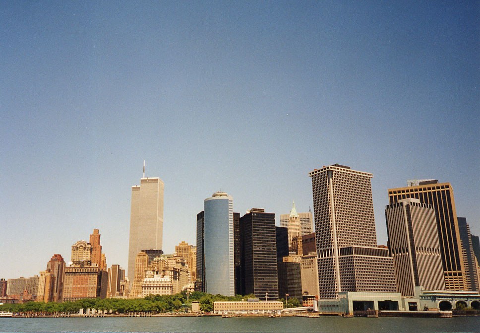 Color photograph of the Lower Manhattan skyline from the water on a clear summer day. The two towers of the World Trade Center rise at the left above the trees of Battery Park; a white cylindrical tower, the dark slabs of the financial district, and a long low pier building along the waterfront fill the rest of the frame under an empty blue sky.
