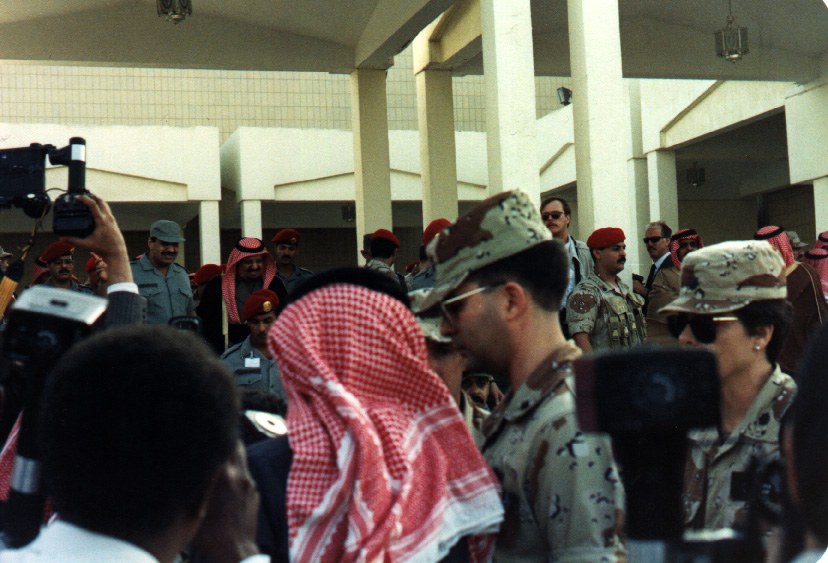 Color photograph, Riyadh Air Base, Saudi Arabia, 1991. A dense crowd packed under a concrete colonnade. In the foreground a television camera is hoisted overhead on a raised arm, lens pointed across the crowd; a second camera body intrudes at the left edge and a third at lower right. Two American officers in desert camouflage and aviator sunglasses stand mid-frame among Saudi officers in red berets and men in checked headdress. Civilians in dark suits and sunglasses behind them. Hard midday shade under the roof slab.