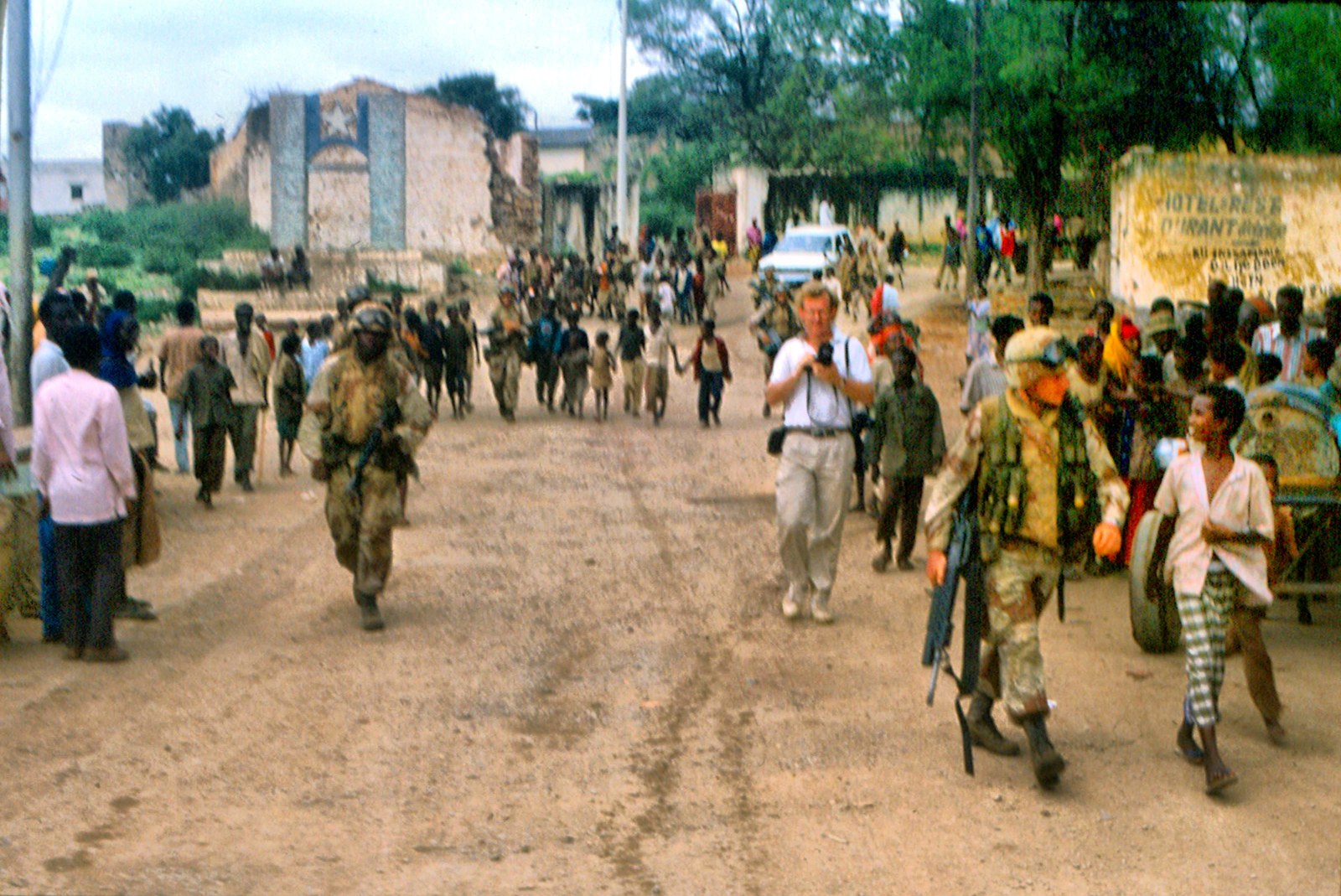 Color photograph, Somalia, 1992. A Western press photographer in a white shirt and pale trousers walks down the middle of a rutted dirt street, two 35mm camera bodies hanging at his chest, one hand steadying a lens. Armed soldiers in camouflage move on either side of him. A crowd of local residents fills both edges of the frame — men, women, children, a handcart with two large wheels at right. Behind them a shelled masonry building with its roof gone, and a wall painted with a star. Overcast light, dust underfoot.