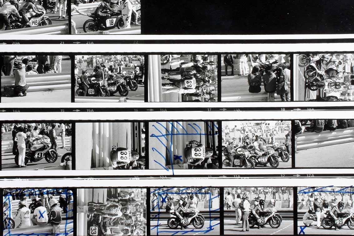 A black-and-white 35mm contact sheet, four strips of frames printed at negative scale with sprocket edges and frame numbers intact. Several frames carry marks made in blue grease pencil: rectangular crop boxes ruled inside two frames, diagonal strokes and X marks struck across others. The photographs on the sheet show a motorcycle-racing paddock — riders, machines, spectators along a barrier.