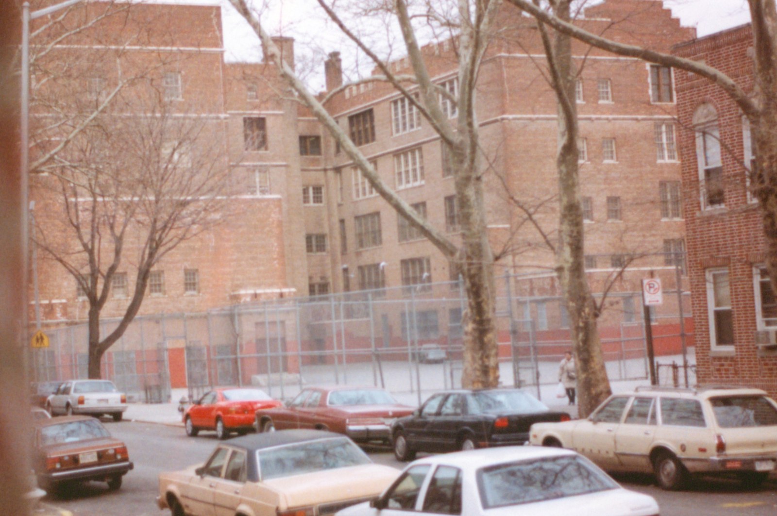 Color photograph of a large brick public school building seen from across a residential street in winter. The school is four and five storeys, plain brick with stone banding and tall multi-pane windows, a curved bay at the centre; a high chain-link fence encloses an empty paved yard along its front, the fence backed by a painted red band on the wall behind. Bare plane trees stand along the kerb, a row of sedans and station wagons is parked at an angle below, and one woman walks past with a shopping bag.