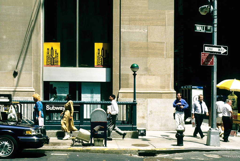 Color photograph of a downtown corner in hard summer sunlight. A green street sign reading WALL ST is bolted high on a lamppost at right, with a one-way arrow and a parking-regulation sign below it. A subway stair with a green globe and an iron railing opens at the kerb beside a mailbox and a fire hydrant; a vendor's yellow-and-white umbrella stands at the frame's edge. Men in white shirts and dark ties walk the sidewalk past a limestone building front, and a dark sedan waits at the corner.