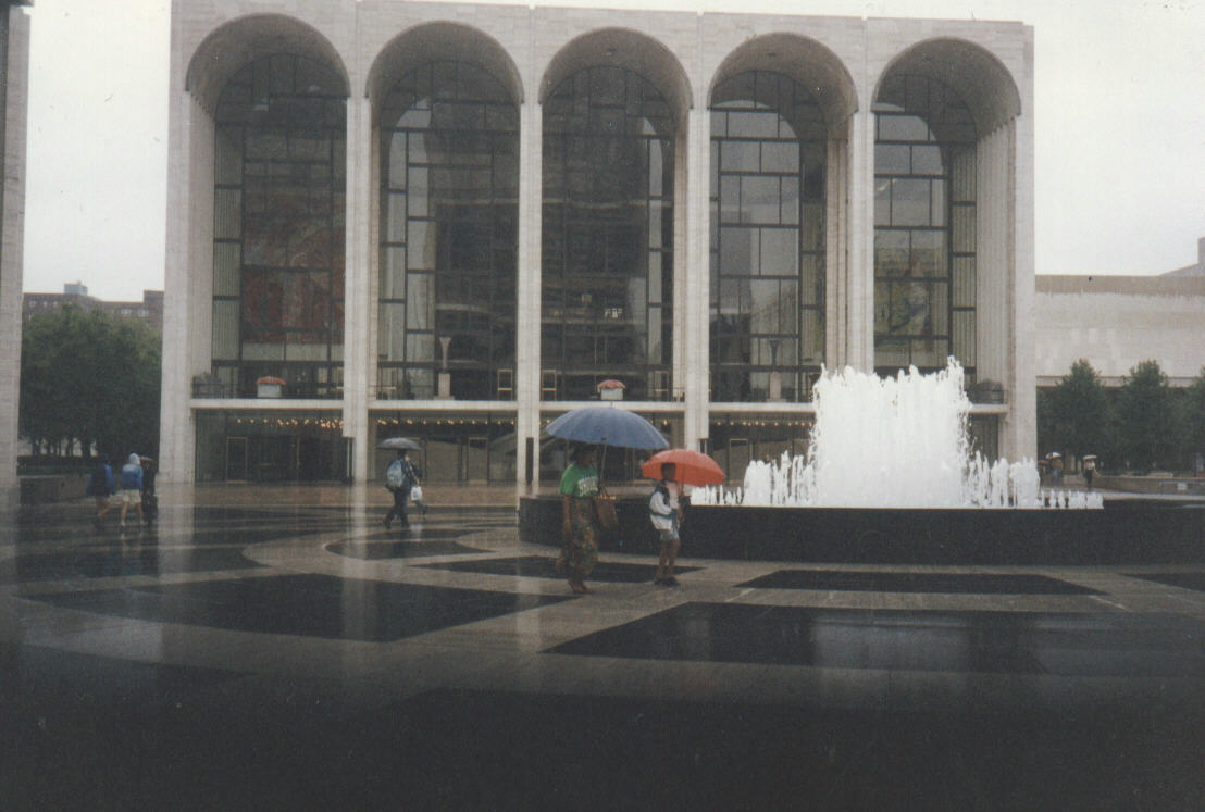 Color photograph of the Metropolitan Opera House seen across a wet plaza. Five tall arches front the travertine facade, glazed from ground to roof, with two large murals visible through the glass behind them. A fountain plays at right; the paving in the foreground is dark with rain and reflects the building. A few figures cross the plaza under umbrellas, one blue and one orange.