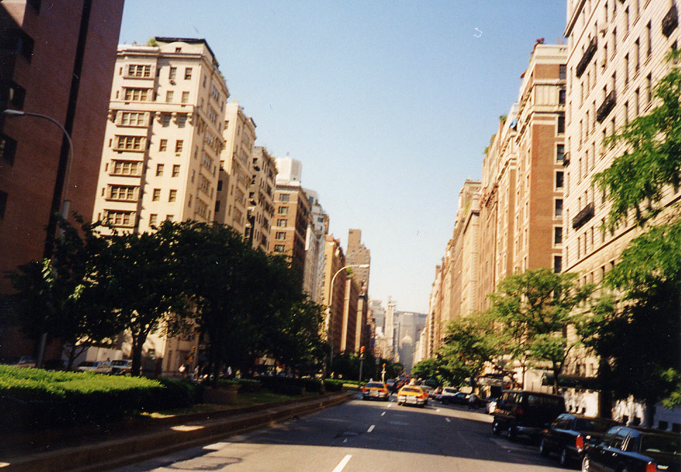 Color photograph looking south along Park Avenue on the Upper East Side. Prewar apartment houses of limestone and dark brick line both sides of the roadway, ten to fifteen storeys, with setbacks, terraces and planted rooftops. A narrow median of clipped hedge and low trees runs down the centre; taxis and dark cars move along the pavement, and a tower closes the view far down the avenue.