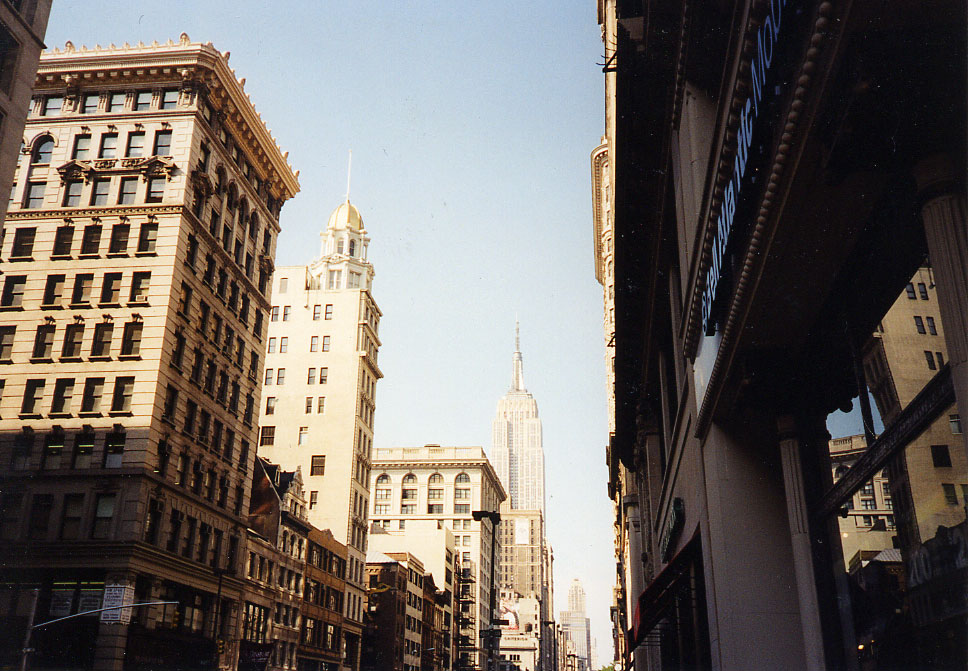 Color photograph looking north along a Manhattan commercial corridor in low late-afternoon sun. Cast-iron and masonry loft buildings of eight to twelve storeys line both sides, heavily corniced, one crowned with a small cupola; awnings and fire escapes hang over the ground floors. The Empire State Building stands at the end of the view down the middle of the street.