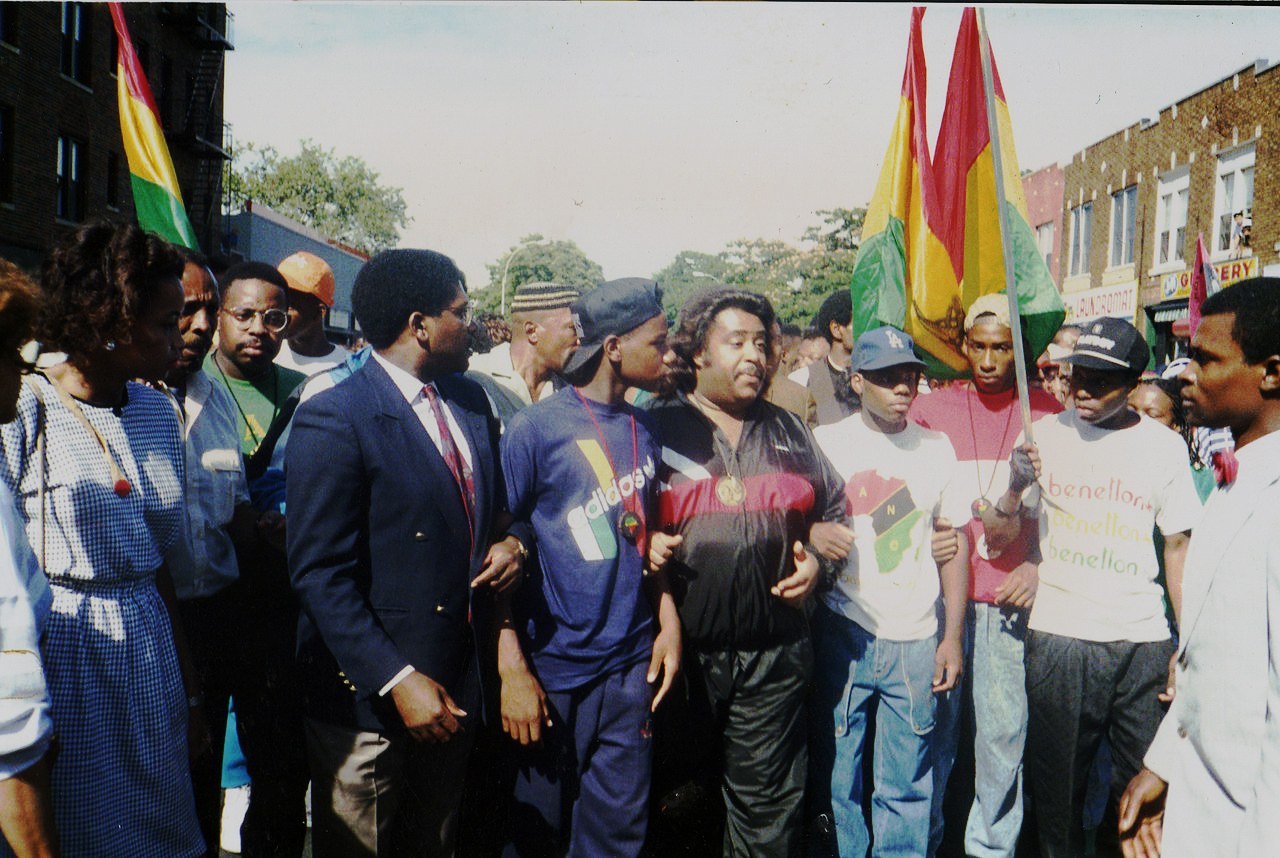 Color photograph of a street demonstration on a bright day. A crowd of Black marchers fills the roadway of a low-rise commercial block, moving toward the camera with arms linked. At the centre a heavyset man in a dark tracksuit and a heavy chain walks between a man in a navy suit and tie and a young man in a blue athletic shirt; teenagers in baseball caps and T-shirts press in behind, two of them carrying large red, gold and green flags on poles. Two-storey brick buildings with shop awnings line the block behind them.