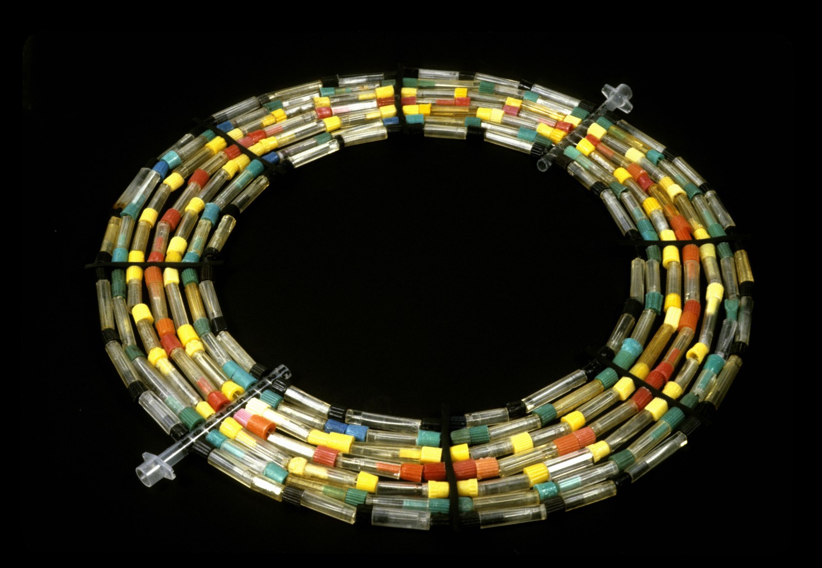 A circular neck collar photographed against black: five concentric strands of clear plastic crack vials strung side by side, punctuated by their snap-on caps in yellow, red, green, blue, and orange, with two clear syringes worked into the strands as fasteners.