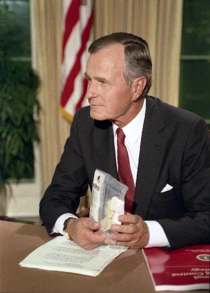 A man in a dark suit and red tie sits at a desk in a formal office, turned in profile toward a camera off to his right, holding up beside his shoulder a small clear plastic evidence bag containing several off-white lumps. Typed speech pages lie open on the desk in front of him alongside a red folder; an American flag and heavy drapes stand behind him.