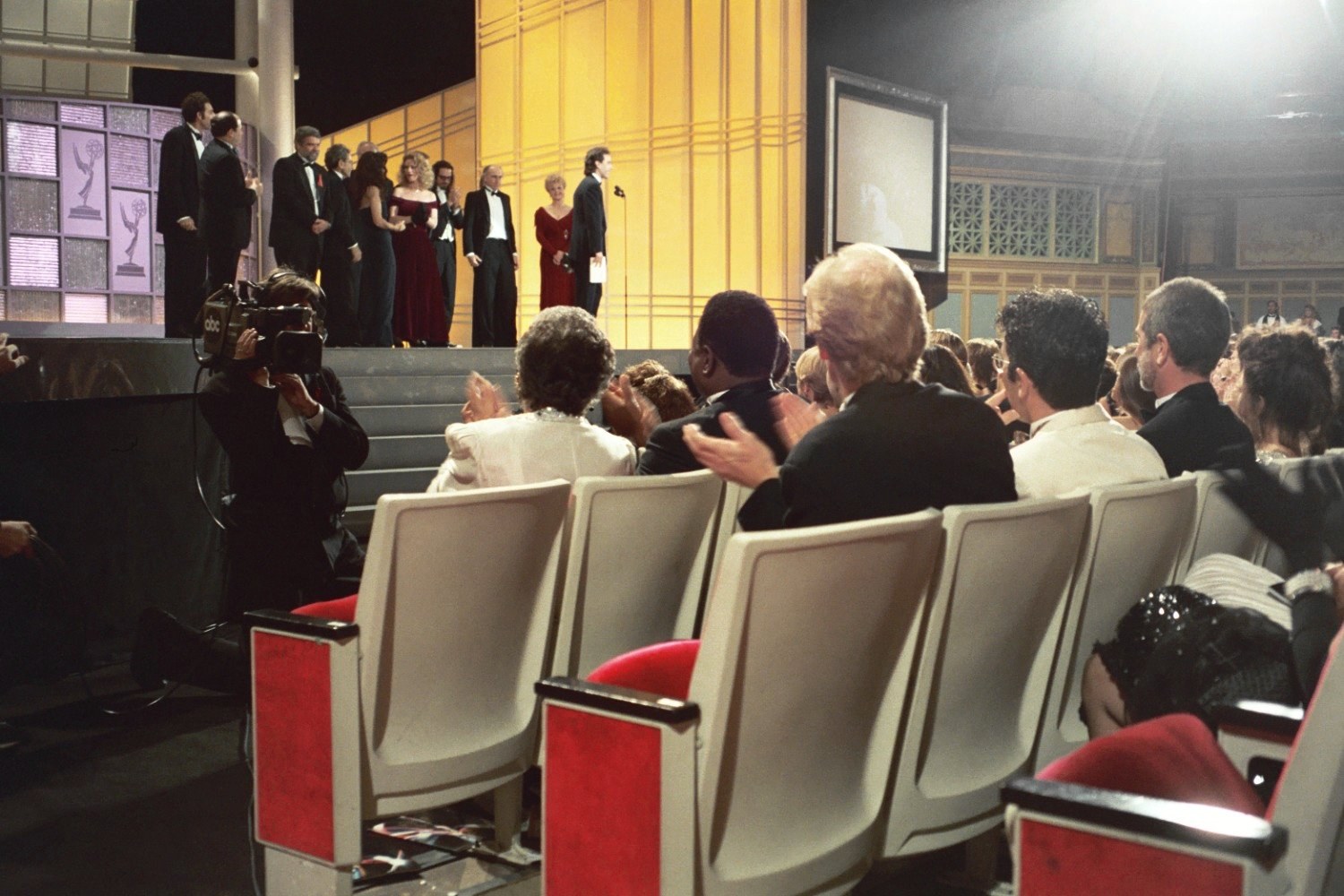A colour photograph taken from within a theatre audience during an awards telecast. In the foreground, the pale curved backs of upholstered seats with red-fronted armrests, several of them empty, and beyond them rows of seated people seen from behind. At the left, a television camera operator crouches at the foot of the stage steps with a shoulder camera trained on the audience. On the lit stage beyond, a line of small distant figures in evening dress stands at a microphone in front of a gold Art Deco backdrop, with a panel of award statuettes to the far left.
