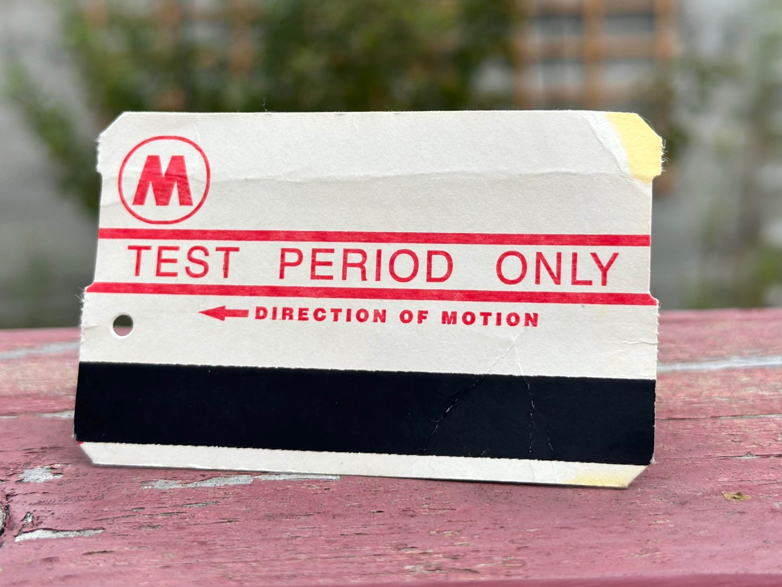 A worn paper fare card standing upright on a weathered plank of pink-painted wood, photographed close and at a slight angle. The card is creased across its middle, torn at two corners, and carries yellow tape residue at the upper and lower right. Printed in red on the white face: a circled M roundel at upper left, then TEST PERIOD ONLY between two heavy rules, then a left-pointing arrow and the words DIRECTION OF MOTION. A punched hole sits at the left edge. A black magnetic stripe runs the full width of the lower third.