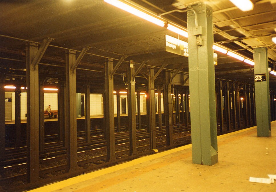 Colour photograph of an empty subway platform, its whole depth washed warm amber by fluorescent tubes running along the ceiling beams. A row of pale green steel columns marches into the distance, the nearest carrying the numerals 23. Black riveted girders and a second row of columns separate the platform from the track bed in the foreground. Across the tracks, a single person sits alone on a bench against white tile under a strip of light. The platform edge runs yellow along the right.