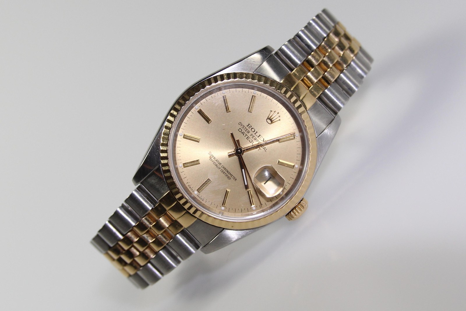 Studio photograph of a wristwatch lying at a three-quarter angle on a plain pale gray ground, its bracelet curving away behind it. The case is steel with a fluted gold bezel, about the width of a man's wrist bone; the dial is champagne-gold with applied baton markers, slim gold hands, and a small date window at the three o'clock position covered by a raised magnifying bubble in the crystal. The bracelet alternates steel and gold links in five rows. The lighting is even and shadowless.