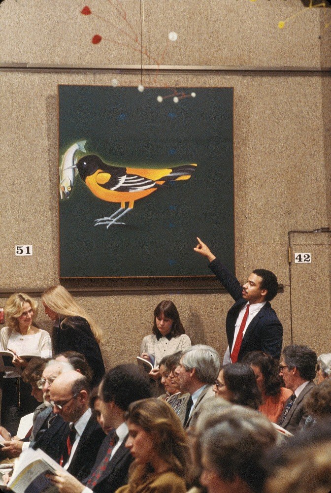 A contemporary-art auction in progress. A young man in a dark blazer and red tie stands at the side of a burlap-panelled saleroom wall with his arm raised, one finger up, beneath a large painting of an oriole holding a fish in its beak against a dark green ground. Numbered lot boards hang on the wall to either side of him. In front of him, rows of seated bidders sit shoulder to shoulder with printed catalogues open in their laps, most of them looking down rather than up.