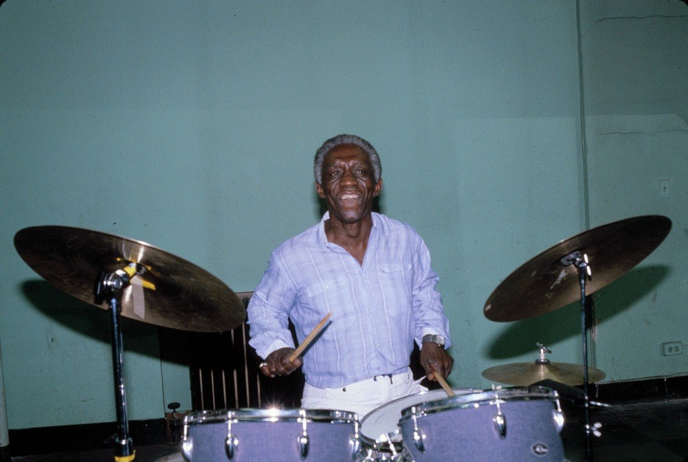 A grey-haired drummer in a pale striped shirt sits grinning behind a drum kit, sticks in both hands, a ride cymbal and a crash cymbal angled toward him on either side and a hi-hat to his right. The room behind him is a bare pale-green wall with a folding screen and an electrical outlet, no stage lighting and no audience.