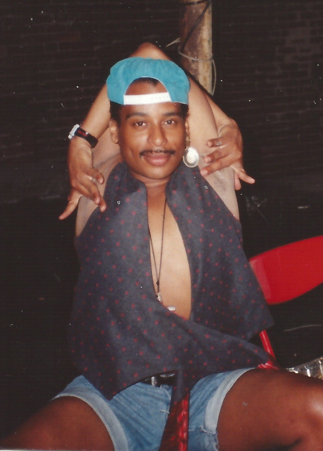 A young man sits in a dark room with an exposed brick wall behind him, wearing a backwards teal baseball cap, a large single pearl earring, and a dark red-dotted shirt hanging open off both shoulders over cut-off denim shorts. His arms are folded up and back over the top of his head, hands framing his face from behind in a held pose, and he is smiling at the camera.