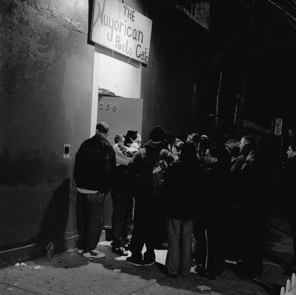 A black-and-white night photograph of a crowd of about twenty people pressed around a lit doorway in an otherwise dark tenement wall. A hand-lettered board above the door reads The Nuyorican Poets Cafe. The door itself carries three metal house numerals. The people wait bunched on the sidewalk in winter coats and hooded jackets, most with their backs to the camera, and litter lies against the wall at the frame's left edge.